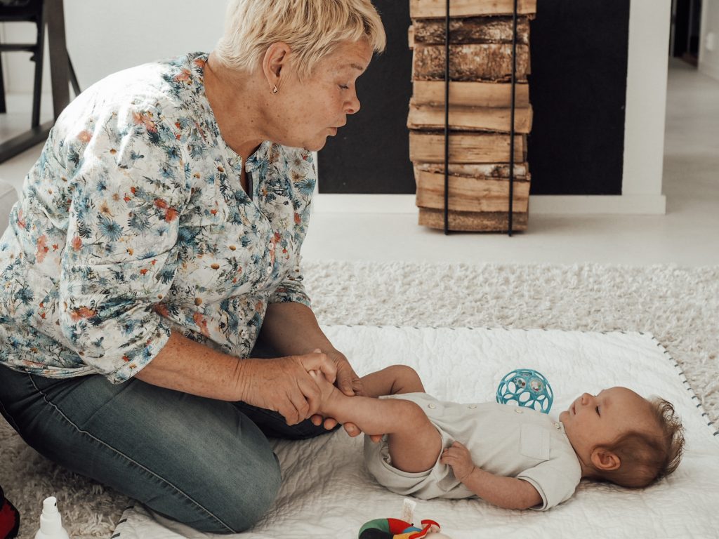 Kuvassa vaaleahiuksinen nainen istuu lattialla ja katsoo lattialla makaavaan vauvaan samalla kun hieroo tämän vasenta jalkaa. Lattialla lapsen vierellä on pallo ja helistinlelu. Taustalla näkyy polttopuuteline sekä keittiön pöytä tuoleineen.