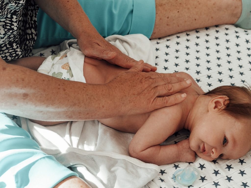 Kuvassa oletettavasti nainen istuu lattialla ja hieroo lattialla makaavaan vauvan selkää. Vauvalla on yllään vain vaippa. Lattialla lapsen vierellä on tutti.